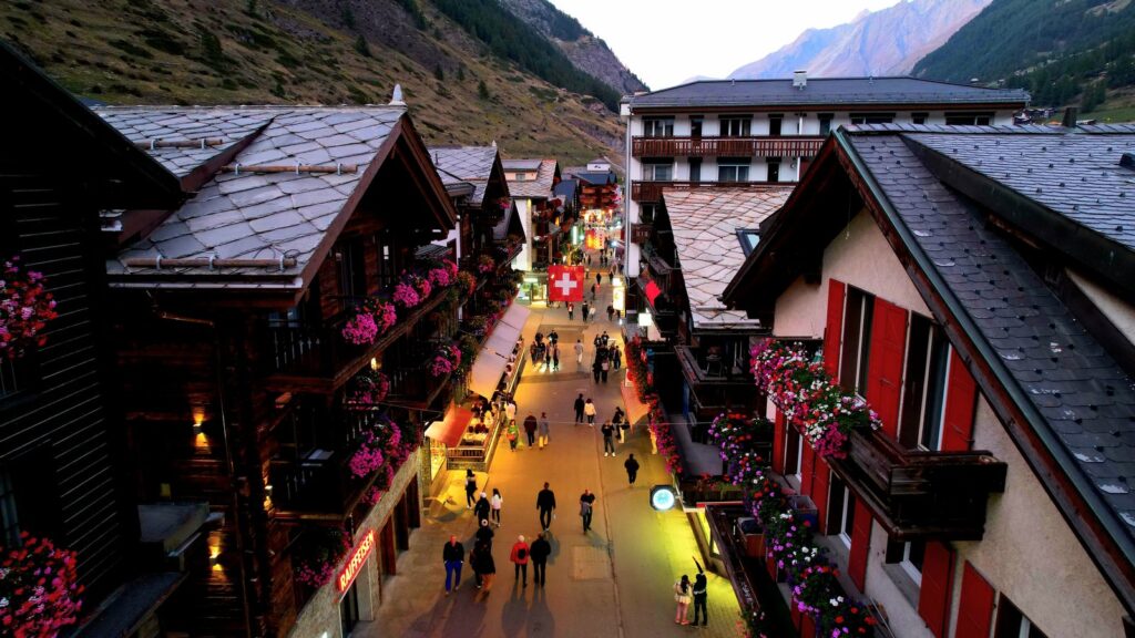 The main street of Zermatt with flower-decorated wooden chalets, Swiss flags, and tourists strolling through the car-free village