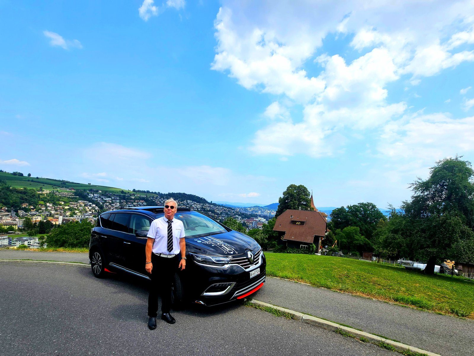 専用車 Renault Espace Initiale の前に立つガイドのTuri。背景に小さな礼拝堂と緑の丘。