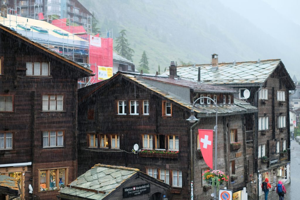 雨に濡れた山あいの村の木造家屋とスイス国旗。斜面に霧がかかっている
